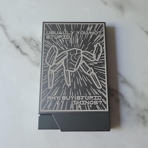 Op de afbeelding: Een rechthoekige donkergrijze metalen kaarthouder met een lasergravure van een gestileerde robot. De tekst USUALLY YOU NOT STUPID staat bovenaan en WHY BUY STUPID THINGS? onderaan, omringd door uitstralende lijnen.
