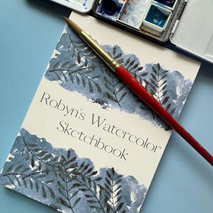 May include: A watercolor sketchbook with the text "Robyn's Watercolor Sketchbook" and a painted blue and black leaf design. A red-handled paintbrush rests on the sketchbook, next to a metal watercolor paint palette with various colors.