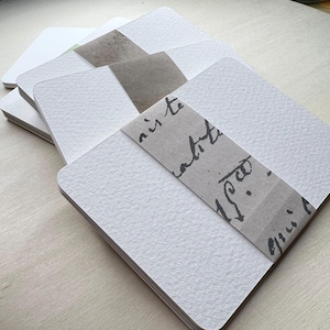 May include: A stack of white textured paper cards with a brown paper band wrapped around them. The band has black handwritten text on it.