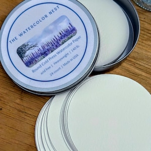 May include: A round metal tin labeled "THE WATERCOLOR NEST" with a watercolor painting of lavender fields. The tin contains round, white watercolor paper. The label indicates the paper is cold press, acid-free, heavyweight, and 140 lb. 24 count.