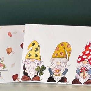 May include: Three gnomes with different colored hats and outfits. The gnome on the left has a green hat with shamrocks, the middle gnome has a pink and yellow floral hat, and the gnome on the right has a red and white polka dot hat. The gnomes are holding a shamrock, a flower, and a mushroom.