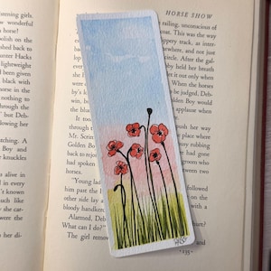 May include: Watercolor bookmark featuring red poppy flowers with green stems and grass against a blue and pink sky. The bookmark is rectangular and has a painted design, perfect for book lovers.