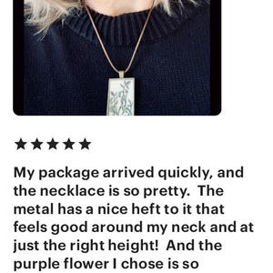 May include: A close-up of a woman wearing a necklace with a purple flower pendant. The necklace is made of metal and has a delicate design. The woman is wearing a black sweater.