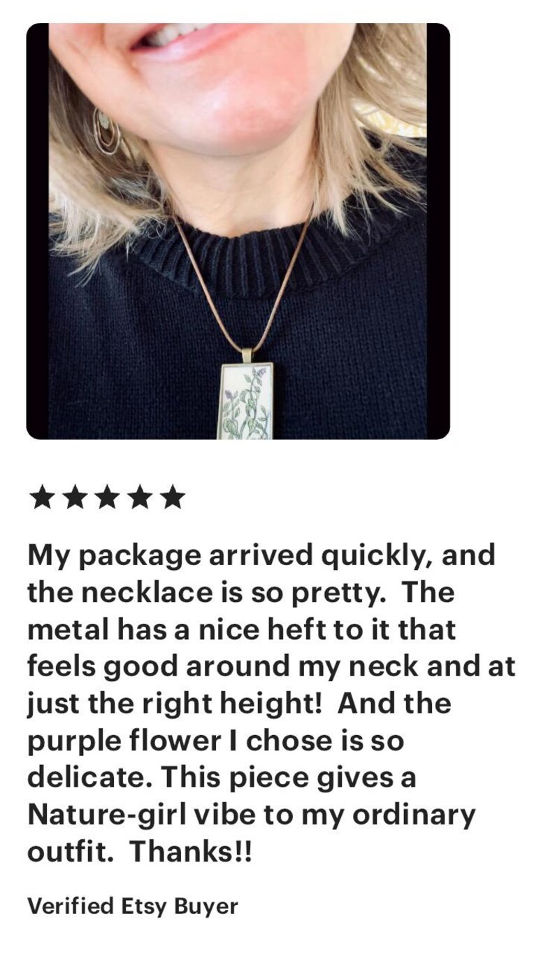 May include: A close-up of a woman wearing a necklace with a purple flower pendant. The necklace is made of metal and has a delicate design. The woman is wearing a black sweater.