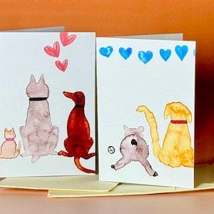 May include: Two greeting cards with watercolor illustrations of dogs and cats. The first card features a white dog, a brown dog, and a cat, all looking up at a row of pink hearts. The second card features a gray cat, a yellow dog, and a blue dog, all looking up at a row of blue hearts.