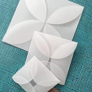 May include: Three white paper envelopes of varying sizes, each with a petal-like design. A U.S. dime is next to the smallest envelope for size comparison. The envelopes are on a teal textured surface.