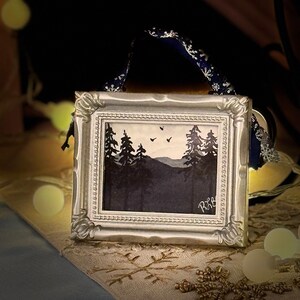 May include: A small, framed watercolor painting of a forest scene with silhouetted trees and mountains. The painting is in a silver-colored ornate frame and has a blue ribbon for hanging. The artwork is signed in the lower right corner.