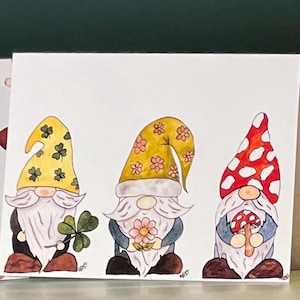 May include: A colourful illustration of three gnomes. The gnomes have white beards and wear hats in yellow and red with white spots. One gnome holds a four-leaf clover, another a flower, and the third a mushroom.