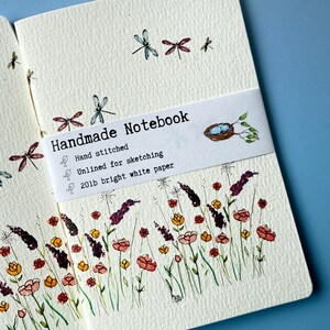 May include: Open handmade notebook with floral and dragonfly illustrations. The notebook features a white band with the text "Handmade Notebook", "Hand stitched", "Unlined for sketching", and "20lb bright white paper". The notebook is decorated with watercolor flowers.