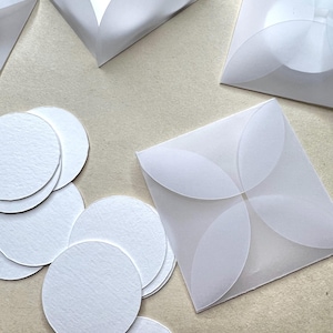 May include: A stack of white round paper circles, some are in a white envelope with a scalloped edge, and others are in a white envelope with a four-petal design.