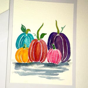 May include: Watercolour painting of five pumpkins in shades of orange, red, teal, pink, and purple. Each pumpkin has a green stem and leaf. The pumpkins are arranged in a row with a light grey shadow beneath them.