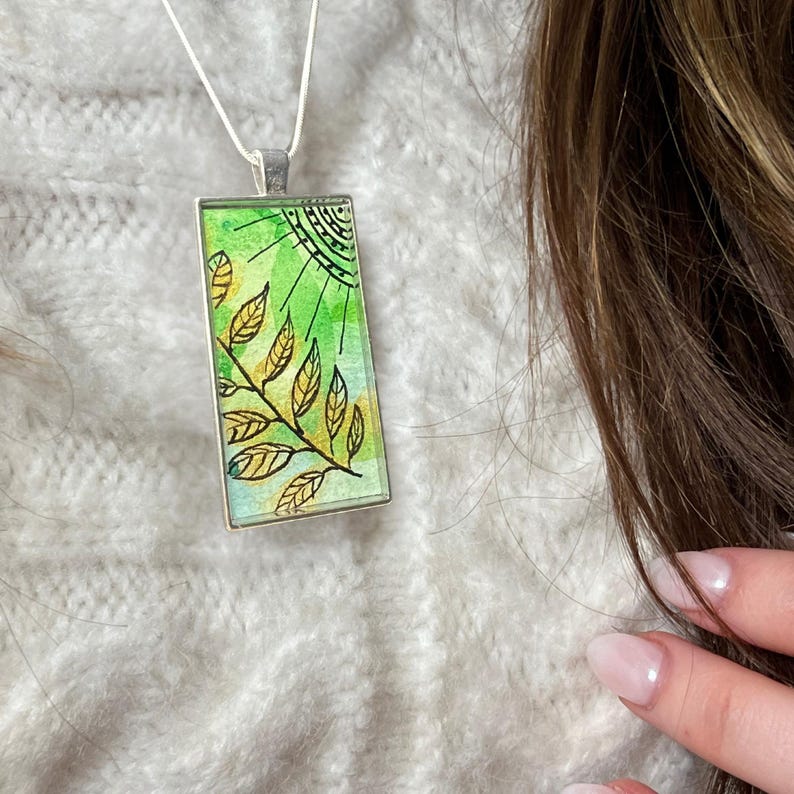 May include: A silver chain necklace with a rectangular pendant featuring a green and gold watercolour design of a sun and leaves.