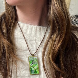 May include: A green and white rectangular pendant with a floral design hangs from a brown cord necklace. The pendant is framed in gold metal.