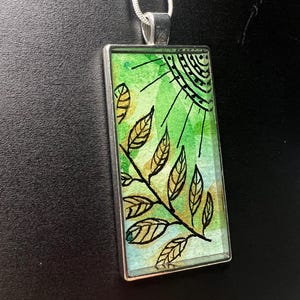 May include: A silver pendant necklace with a rectangular glass charm featuring a green and blue watercolour background with a black and gold leaf design.