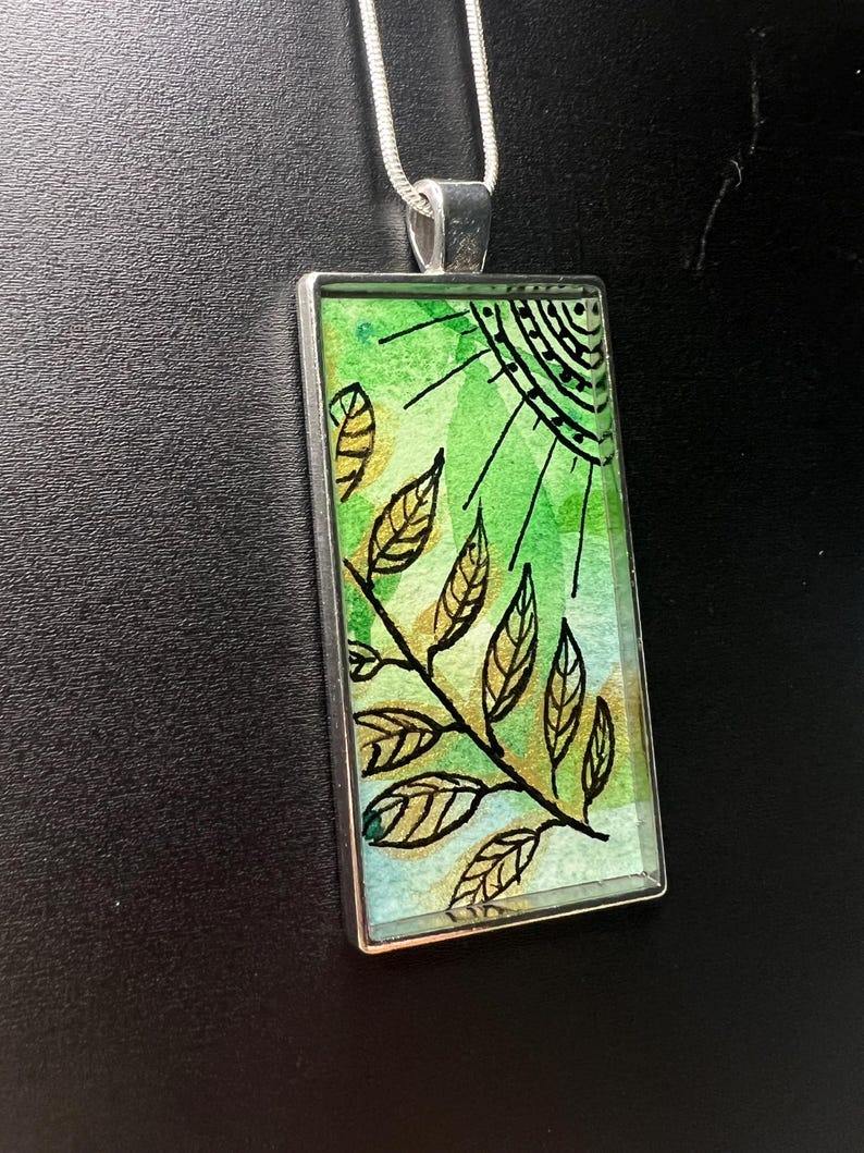 May include: A silver pendant necklace with a rectangular glass charm featuring a green and blue watercolour background with a black and gold leaf design.