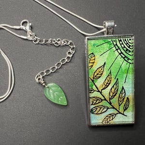May include: A silver chain necklace with a green and gold rectangular pendant featuring a watercolour-style design of leaves and a sun. The pendant is attached to a silver chain with a green jade leaf charm.