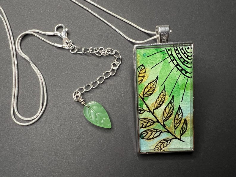 May include: A silver chain necklace with a green and gold rectangular pendant featuring a watercolour-style design of leaves and a sun. The pendant is attached to a silver chain with a green jade leaf charm.