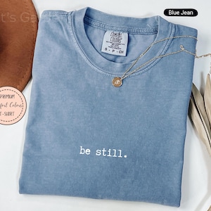 Könnte beinhalten: Ein blaues Comfort Colors T-Shirt mit dem Aufdruck "be still" auf der Vorderseite. Eine goldene Halskette mit Anhänger liegt auf dem Shirt. Das Shirt ist auf einer weißen Oberfläche gefaltet.