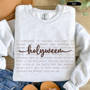 K&ouml;nnte beinhalten: Hellgraues Sweatshirt mit dem Wort "holyween" in Schreibschrift. Das Sweatshirt hat einen Rundhalsausschnitt und zeigt ein Gedicht &uuml;ber Halloween. Der Text ist braun.