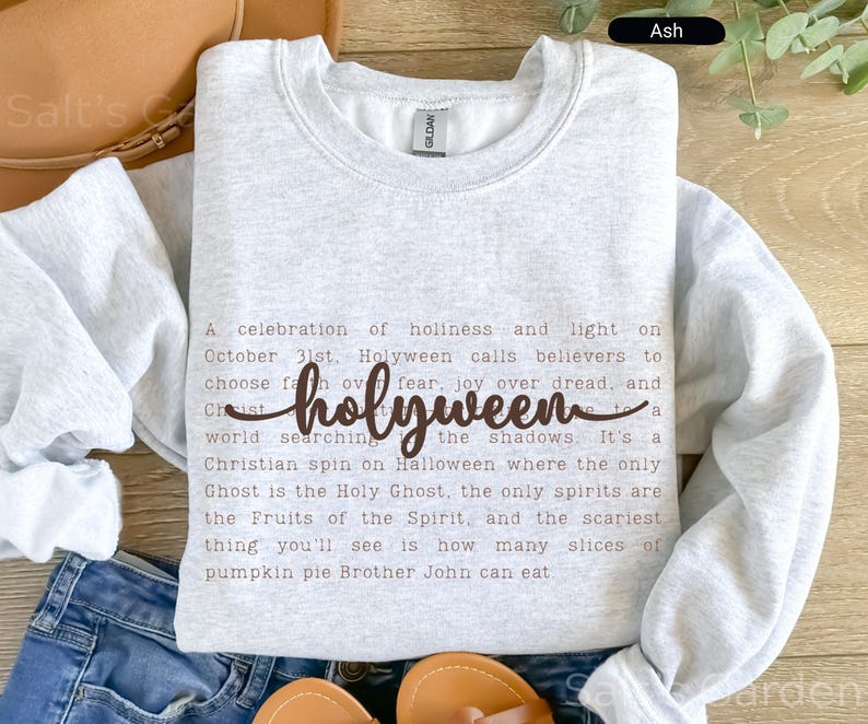 K&ouml;nnte beinhalten: Hellgraues Sweatshirt mit dem Wort "holyween" in Schreibschrift. Das Sweatshirt hat einen Rundhalsausschnitt und zeigt ein Gedicht &uuml;ber Halloween. Der Text ist braun.