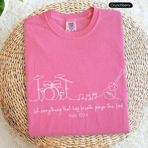 May include: A pink t-shirt with a white line drawing of a drum set, guitar, and musical notes. The text "Let everything that has breath praise the Lord Psalm 150:6" is printed below the drawing. The shirt is on a woven surface.