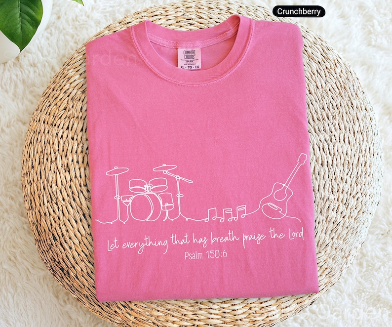 May include: A pink t-shirt with a white line drawing of a drum set, guitar, and musical notes. The text "Let everything that has breath praise the Lord Psalm 150:6" is printed below the drawing. The shirt is on a woven surface.