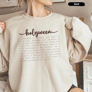 May include: Sand-colored sweatshirt with the word "holyween" in a script font. Below the word is a paragraph of text. The sweatshirt is a neutral color and has long sleeves.