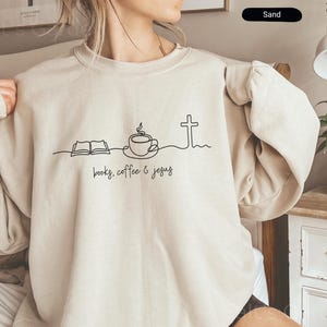 May include: Sand-colored sweatshirt with a crew neck. The front features a minimalist black line drawing of an open book, a coffee cup, and a cross, with the words "books, coffee & jesus" below.