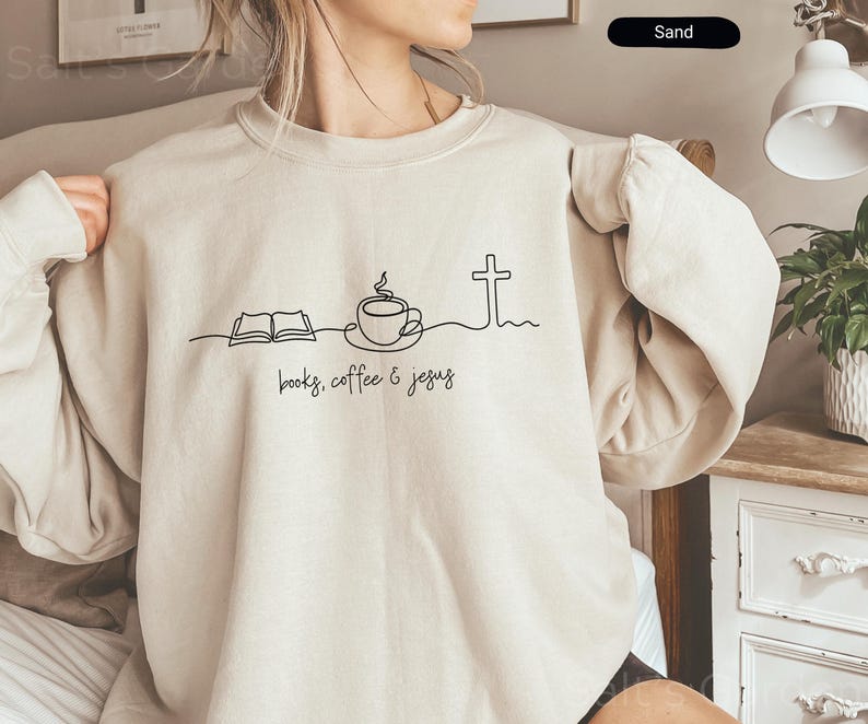 May include: Sand-colored sweatshirt with a crew neck. The front features a minimalist black line drawing of an open book, a coffee cup, and a cross, with the words "books, coffee & jesus" below.