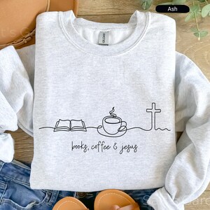 May include: Ash gray sweatshirt with a black line drawing of an open book, coffee cup, and cross. The words "books, coffee & jesus" are written below the drawing. The sweatshirt is made of a soft material.