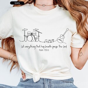 May include: Ivory-colored t-shirt featuring a minimalist line drawing of a drum set, musical notes, and a guitar. The text "Let everything that has breath praise the Lord Psalm 150:6" is printed below the graphic.