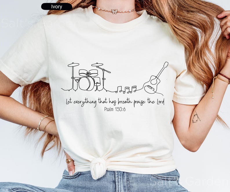 May include: Ivory-colored t-shirt featuring a minimalist line drawing of a drum set, musical notes, and a guitar. The text "Let everything that has breath praise the Lord Psalm 150:6" is printed below the graphic.