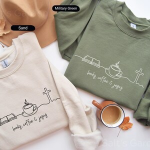 May include: Two crewneck sweatshirts, one sand-colored and one military green, are displayed. Each features a minimalist line drawing of an open book, coffee cup, and cross, with the text "books, coffee & jesus" in a script font. A hat and a mug are also visible.