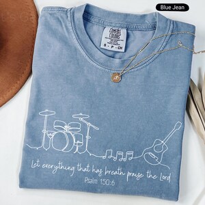 May include: Blue jean colored t-shirt with a white line drawing of a drum set, guitar, and musical notes. The text "Let everything that has breath praise the Lord Psalm 150:6" is printed in white. A gold necklace is draped across the shirt.