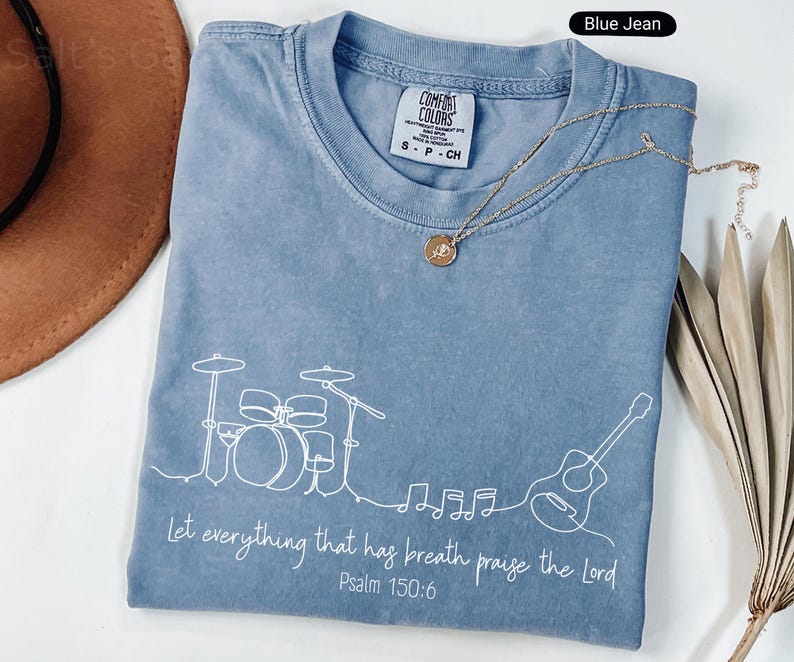 May include: Blue jean colored t-shirt with a white line drawing of a drum set, guitar, and musical notes. The text "Let everything that has breath praise the Lord Psalm 150:6" is printed in white. A gold necklace is draped across the shirt.