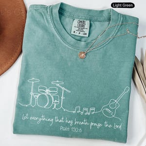 May include: Light green Comfort Colors t-shirt with a white line drawing of a drum set, guitar, and musical notes. The text "Let everything that has breath praise the Lord Psalm 150:6" is printed in white. A gold necklace is draped across the shirt.