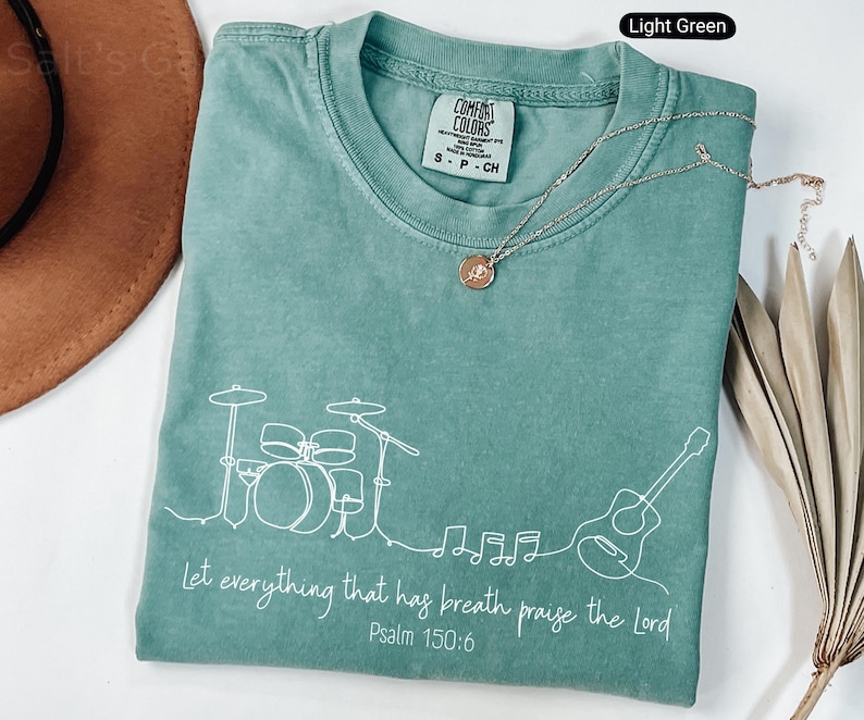 May include: Light green Comfort Colors t-shirt with a white line drawing of a drum set, guitar, and musical notes. The text "Let everything that has breath praise the Lord Psalm 150:6" is printed in white. A gold necklace is draped across the shirt.