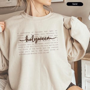 K&ouml;nnte beinhalten: Ein sandfarbenes Sweatshirt mit dem Wort "holyween" in einer Schreibschrift. Das Sweatshirt hat einen Rundhalsausschnitt und lange &Auml;rmel. Der Text auf dem Shirt beschreibt eine christliche Variante von Halloween.