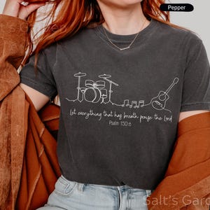 May include: Dark gray t-shirt with a white line drawing of a drum set, musical notes, and a guitar. The text "Let everything that has breath praise the Lord Psalm 150:6" is printed below the drawing. The shirt is a Pepper color.