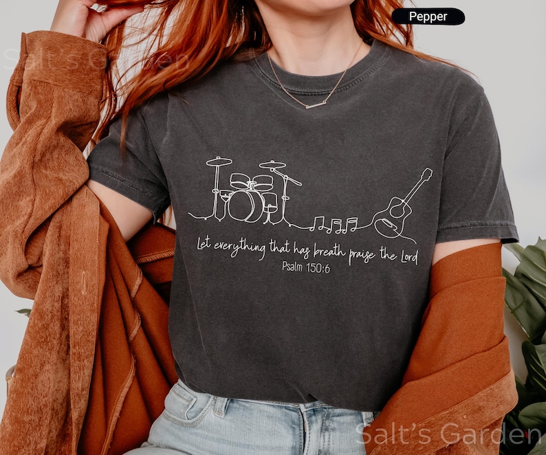 May include: Dark gray t-shirt with a white line drawing of a drum set, musical notes, and a guitar. The text "Let everything that has breath praise the Lord Psalm 150:6" is printed below the drawing. The shirt is a Pepper color.