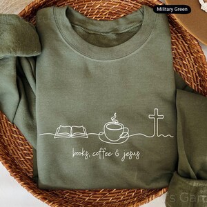 May include: Military green sweatshirt with white line art of an open book, coffee cup, and cross. The words "books, coffee & jesus" are printed below the graphic. The sweatshirt is folded in a woven basket.