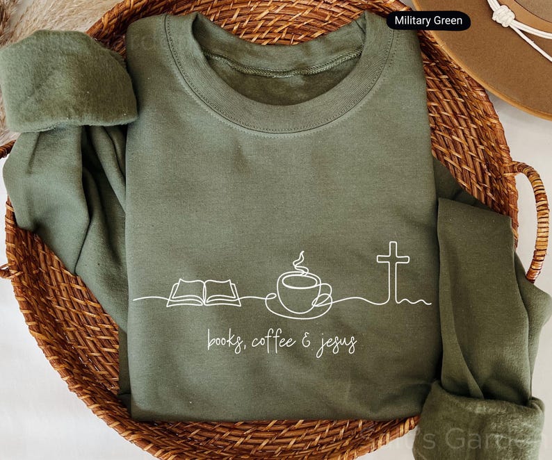 May include: Military green sweatshirt with white line art of an open book, coffee cup, and cross. The words "books, coffee & jesus" are printed below the graphic. The sweatshirt is folded in a woven basket.