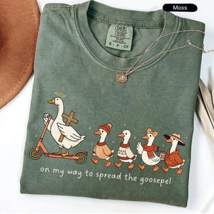 以下が含まれることがあります： モスグリーンのComfort Colors Tシャツ。スクーターに乗ったり歩いたりするガチョウのグラフィック。ガチョウは帽子やスカーフを身につけ、コーヒーカップを持っています。テキストは「on my way to spread the goosepel」と書かれています。