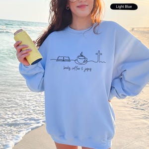 May include: Light blue sweatshirt with a black line drawing of a book, coffee cup, and cross, with the words "books, coffee & jesus" printed below. A yellow can cooler is held in the hand.