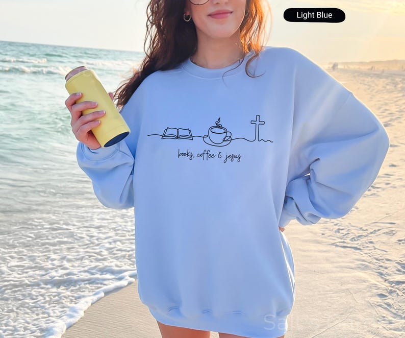 May include: Light blue sweatshirt with a black line drawing of a book, coffee cup, and cross, with the words "books, coffee & jesus" printed below. A yellow can cooler is held in the hand.