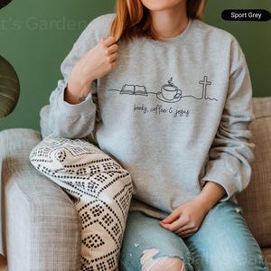 May include: Sport Grey sweatshirt with a black line drawing of an open book, coffee cup, and cross. The words "books, coffee & jesus" are written below the drawing. The model is wearing blue jeans.