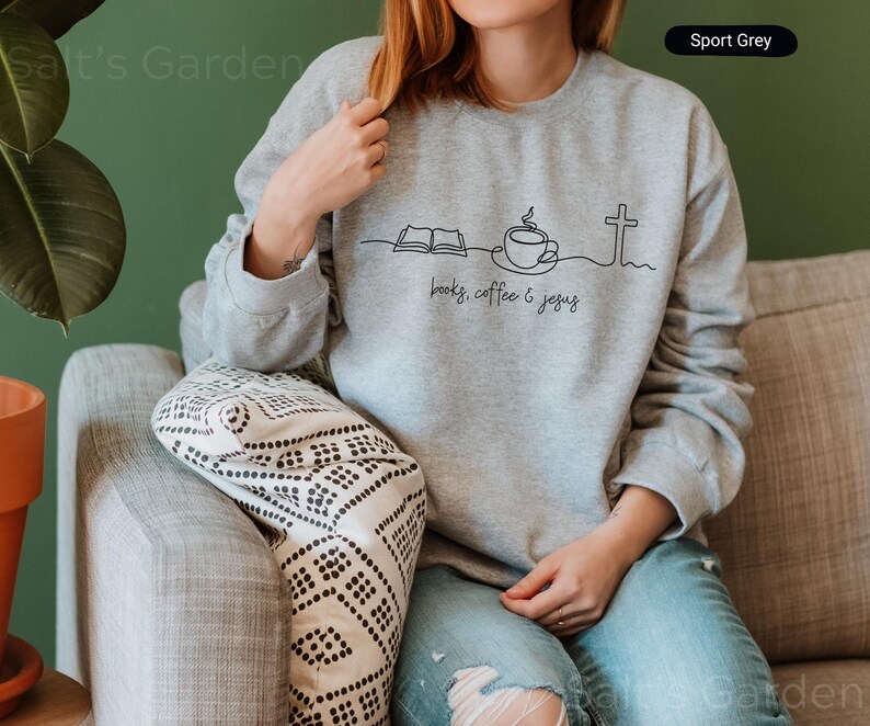 May include: Sport Grey sweatshirt with a black line drawing of an open book, coffee cup, and cross. The words "books, coffee & jesus" are written below the drawing. The model is wearing blue jeans.
