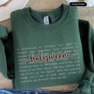 K&ouml;nnte beinhalten: Forstgr&uuml;nes Sweatshirt mit dem Wort "holyween" in Schreibschrift. Das Sweatshirt hat einen Rundhalsausschnitt und lange &Auml;rmel. Der Text auf dem Sweatshirt beschreibt eine christliche Sicht auf Halloween. Das Sweatshirt ist aus weichem Material.
