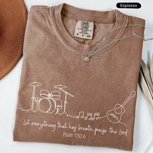 May include: Espresso-colored t-shirt with a white line drawing of a drum set, guitar, and musical notes. The text "Let everything that has breath praise the Lord Psalm 150:6" is printed below the drawing. A silver necklace is draped across the shirt.