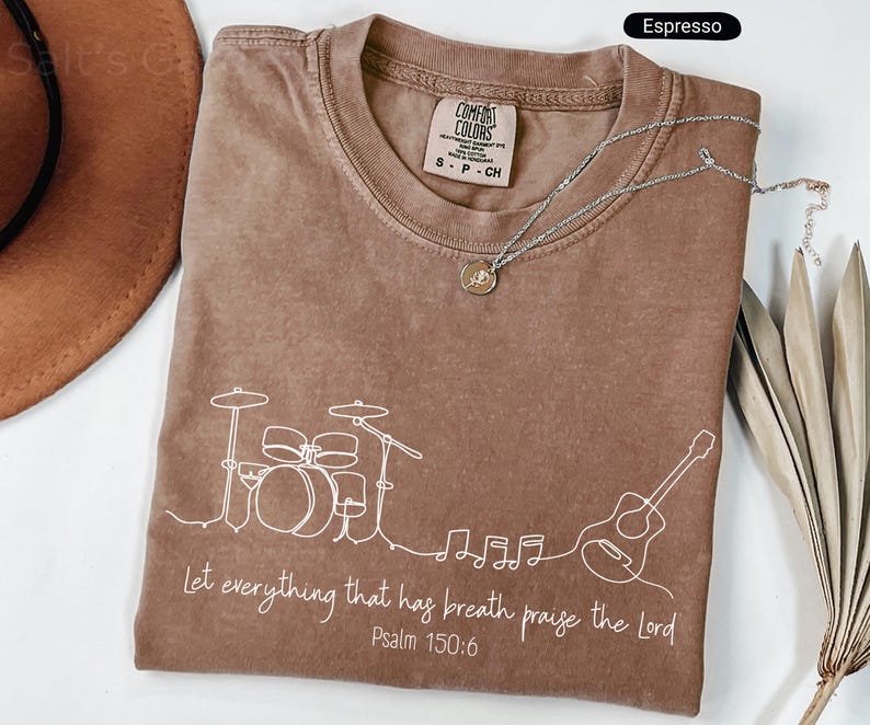 May include: Espresso-colored t-shirt with a white line drawing of a drum set, guitar, and musical notes. The text "Let everything that has breath praise the Lord Psalm 150:6" is printed below the drawing. A silver necklace is draped across the shirt.